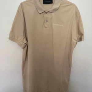 Beige pikétröja från Peak Performance - Säljer en stilren beige pikétröja från Peak Performance. Tröjan har en klassisk krage med knappar och korta ärmar. Perfekt för en avslappnad men ändå snygg look. Använd 2 gånger