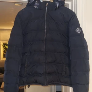 Svart dunjacka från Gant - Säljer en stilren blå dunjacka från Gant med huva och dragkedja. Jackan har ett diskret märke på ärmen och Gant-logga längs dragkedjan. Perfekt för kyliga dagar.