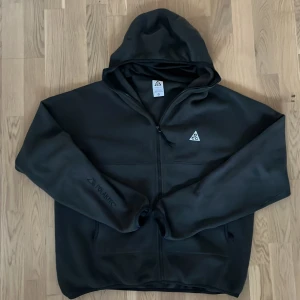 Svart fleecejacka från Nike ACG - Säljer en svart fleecejacka från Nike ACG med huva och dragkedja. Jackan har en enkel design med ACG-logga på bröstet och är perfekt för kyliga dagar. Den har långa ärmar och är tillverkad i mjukt fleece-material. Den har också inbyggd balaclava. Strl L
