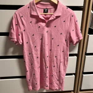 Rosa pikétröja med små svarta mönster - Snygg rosa pikétröja från FSBN med små svarta mönster över hela tyget. Tröjan har en klassisk krage och korta ärmar, perfekt för en avslappnad stil. Passar bra till både jeans och shorts.