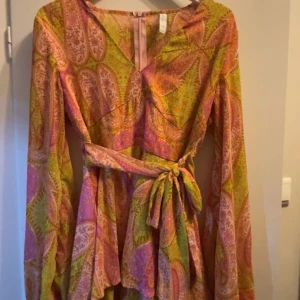 Färgglad mönstrad klänning med knytband - Säljer en färgglad klänning med ett livligt mönster i rosa, grönt och orange. Klänningen har långa ärmar och ett knytband i midjan för en fin passform. Perfekt för sommarn. Den är i storlek M men passar perfekt för mig som är S. Den har även inbyggda shorts under. Skriv för fler bilder💞