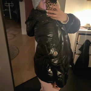 Svart dunjacka från Moncler - Säljer en snygg svart dunjacka från Moncler. Jackan har en glansig yta och en stor huva för extra värme. Perfekt för kyliga dagar. Den har en dragkedja framtill och Monclers logotyp på sidan av armen.