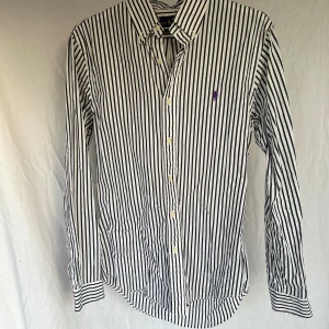 Randig skjorta från Ralph Lauren - Snygg vit och svart randig skjorta från Ralph Lauren med klassisk krage och broderad logga i lila på bröstet. Skjortan har långa ärmar och knappar framtill. Perfekt för en stilren look!