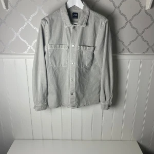 Grå jeansjacka från Zara - Snygg grå jeansjacka från Zara med knappar framtill och två bröstfickor. Perfekt för en avslappnad stil. Jackan har långa ärmar och en klassisk krage.