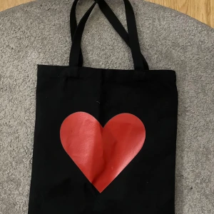 Svart shoppingväska med rött hjärta - Säljer en svart shoppingväska med ett stort rött hjärta på framsidan. Väskan har långa handtag och är perfekt för att bära med sig det nödvändigaste. Enkel och stilren design som passar till många tillfällen.