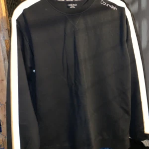 Svart långärmad tröja från Calvin Klein - Snygg svart långärmad tröja från Calvin Klein med vita ränder längs ärmarna. Tröjan har en rund halsringning och Calvin Klein-loggan tryckt på bröstet och insidan av kragen. Perfekt för en stilren och avslappnad look.