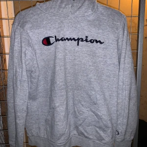 Grå hoodie från Champion - Säljer en grå hoodie från Champion med deras ikoniska logga broderad på bröstet. Tröjan har en klassisk design med huva och långa ärmar. Perfekt för en avslappnad stil. Litet märke på armen, möjligen går det bort när den tvättas. Barnstorlek 