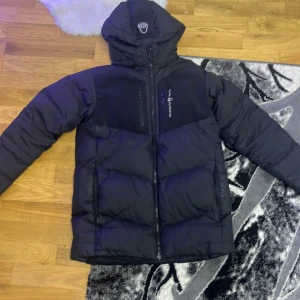 Svart dunjacka från Sail Antarctica Expedition - Säljer en svart dunjacka från Sail Antarctica Expedition. Jackan har en dragkedja framtill och en hög krage för extra värme. Perfekt för kalla dagar med sin pufferdesign.