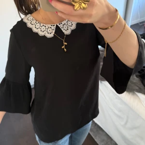 Svart blus med spetskrage - Säljer en elegant svart blus med vacker vit spetskrage. Blusen har trekvartsärmar med volangdetaljer som ger en stilfull touch. Perfekt för att lyfta vilken outfit som helst.