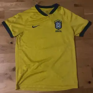 Säljer en gul Brasilien tröja från Nike. Tröjan har korta ärmar och ett broderat Brasilien-emblem på bröstet. Perfekt för fotbollsfans som vill visa sitt stöd för laget! Den är i storlek M