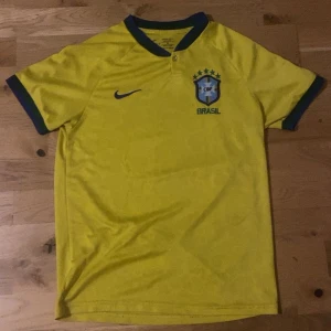 Brasilien tröja  - Säljer en gul Brasilien tröja från Nike. Tröjan har korta ärmar och ett broderat Brasilien-emblem på bröstet. Perfekt för fotbollsfans som vill visa sitt stöd för laget! Den är i storlek M