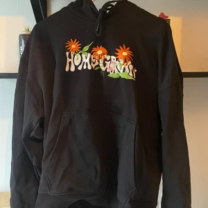 Svart hoodie med broderade blommor från Size? - Säljer en svart hoodie från Size? med coolt broderi av orange blommor och texten 'Homegrown' på framsidan. Den har en klassisk känguruficka och justerbar huva med snören. Perfekt för en avslappnad stil! Använd fåtal gånger därmed nästintill nyskick! 