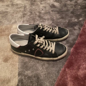 Svarta Phillipe Model med vita detaljer - Snygga svarta sneakers från Philippe Model med vita och röda detaljer. Skorna har en klassisk snörning och en vit sula som ger en stilren look.  Skorna köptes in sommarn 2024