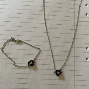 Smyckeset med svart blomma - Elegant smyckeset bestående av ett halsband och ett armband. Båda smyckena har en svart blomma med en glittrig detalj i mitten. Kedjorna är i silverfärg och ger en stilren look. Perfekt för att komplettera din outfit med en subtil men chic touch.