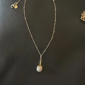 Silverhalsband med vit pärla - Elegant silverhalsband med en vit pärla som hänge. Kedjan är tunn och dekorerad med små detaljer. Hänget har en unik design med en guldliknande infattning runt pärlan. Perfekt för att ge en stilren touch till din outfit.