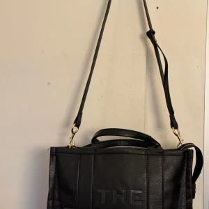 Svart Toteväska. - Snygg svart axelväska från med texten 'THE TOTE BAG' på framsidan. Väskan är tillverkad i skinn och har både handtag och en justerbar axelrem. Perfekt för att bära med sig allt man behöver under dagen. Ej kartongen. 