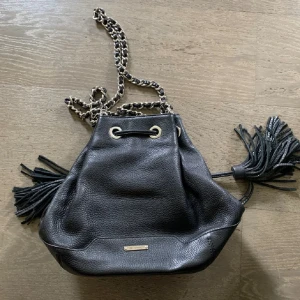 Svart axelväska från Rebecca Minkoff - Snygg svart axelväska i skinn från Rebecca Minkoff med kedjedetaljer och fransar. Väskan har en praktisk dragskoöppning och ett stilrent mönstrat foder. Perfekt för att ge din outfit en edgy touch.
