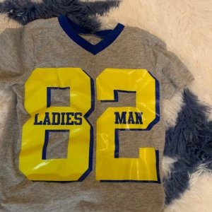 Grå t-shirt med tryck från Okie Dokie - Säljer en grå t-shirt från Okie Dokie med ett stort gult tryck '82 LADIES MAN' på framsidan. T-shirten har en blå v-ringad krage och är kortärmad. Perfekt för en cool och avslappnad stil. Passar 3-4 åring.