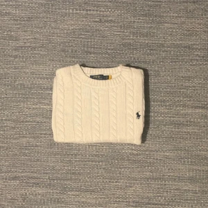 Vit stickad tröja från Ralph Lauren - Säljer en klassisk vit stickad tröja från Ralph Lauren med kabelstickat mönster. Tröjan har långa ärmar och en broderad logotyp på bröstet. Perfekt för en stilren look.