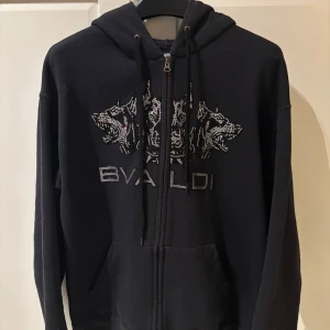 Svart hoodie med dragkedja från Bvaldi - Snygg svart hoodie med dragkedja från Bvaldi. Den har ett coolt mönster med glittriga detaljer på framsidan. Knappt använd har mest hängt i garderoben. Nypris (1100) 