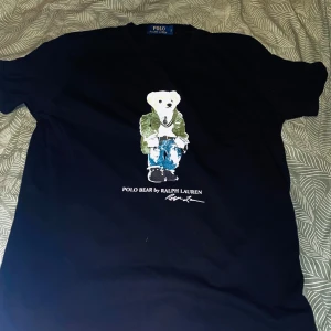 Svart t-shirt med Polo Bear från Ralph Lauren - Snygg svart t-shirt från Ralph Lauren med det ikoniska Polo Bear-trycket på framsidan. Perfekt för en casual look med en touch av stil. Den svart är i Bra skick, däremot har den beigefärgade är i Nytt skick då den har knappast använts. Säljer båda tillsammans för 1000. Passar som M!!