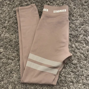 Beiga leggings från Stronger - Snygga bruna leggings från Stronger med hög midja och elastisk resår. De har vita detaljer i form av ränder och prickar på benen. Perfekta för träning eller en avslappnad dag. Inga defekter 