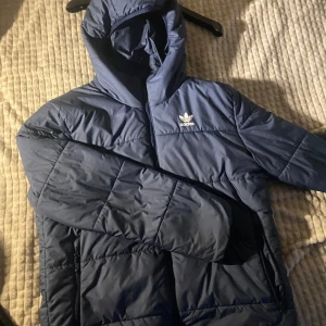 Blå pufferjacka från Adidas - Säljer en blå pufferjacka från Adidas med huva och dragkedja. Jackan har ett quiltat mönster och Adidas-loggan på bröstet. Perfekt för kyliga dagar.