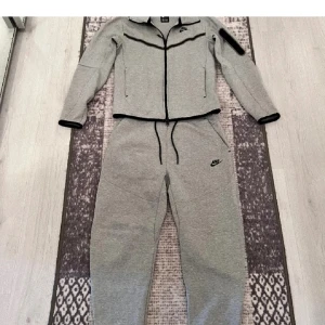 Grå träningsdräkt från Nike - Säljer en grå träningsdräkt från Nike med dragkedja och huva. Dräkten har svarta detaljer och elastiska muddar. Perfekt för träning eller avslappnade dagar.