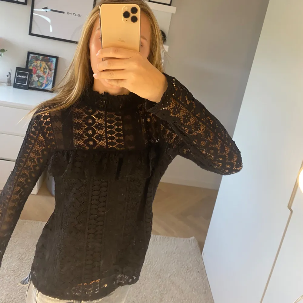 Elegant svart spetsblus med långa ärmar och vackra detaljer. Perfekt för en stilfull look med sitt genombrutna mönster och volangdetalj framtill.. Yläosat.