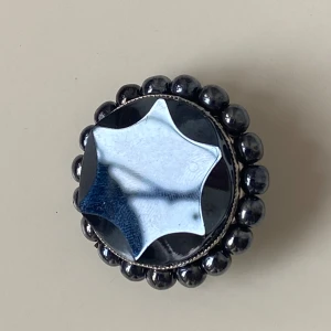 Svart brosch med rund design - Elegant vintage svart brosch med en rund form och en stjärnliknande detalj i mitten. Omgiven av glänsande pärlor som ger en sofistikerad look. Perfekt för att ge en extra touch till din outfit.