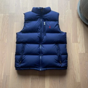 Blå dunväst från Polo Ralph Lauren - Säljer en snygg blå dunväst från Polo Ralph Lauren. Västen har en dragkedja framtill och två praktiska fickor med dragkedjor. Perfekt för kyligare dagar.
