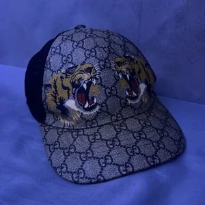 Gucci keps med tigerbroderi - Snygg keps från Gucci med ikoniskt mönster och broderade tigrar. Kepsen går att justera, storleken är S motsvarar 57cm bredd på huvudet.