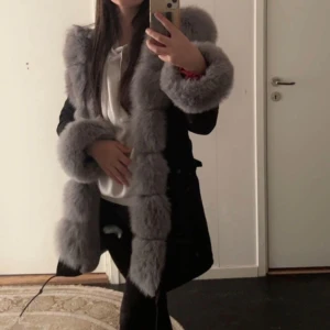 Svart jacka  med grå pälsdetaljer - Snygg svart kappa med lyxiga grå pälsdetaljer längs kragen, ärmsluten och framkanten. Perfekt för att hålla sig varm och stilren under kyliga dagar. Den är köpt för 4000 