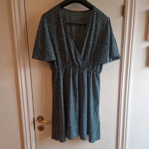 Mörkgrön prickig klänning - Charmig mörkgrön klänning med vita prickar och v-ringning. Klänningen har korta ärmar och en elastisk midja som ger en smickrande passform. Perfekt för en avslappnad och stilfull look.