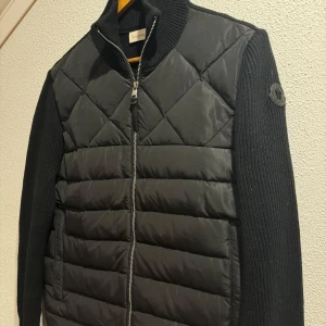 Moncler Cardigan - Snygg svart quiltad jacka från Moncler med dragkedja och ribbad krage. Jackan har långa ärmar och en stilren design med Moncler-logga på ärmen. Perfekt för kyligare dagar.