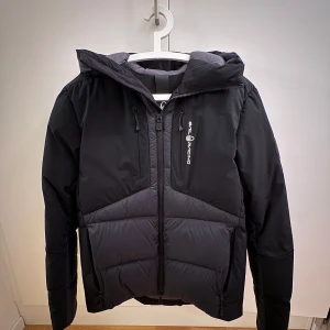 (Använd cirka 10 gånger)Svart vinterjacka från Sail Racing  - Snygg svart dunjacka från Sail Racing med huva och dragkedja. Jackan har en quiltad design och är perfekt för kyliga dagar. Den har en logotyp på bröstet och en på ärmen. Insidan är grå och jackan har flera praktiska fickor. Storlek XS men passar även S. Nypris 5000kr