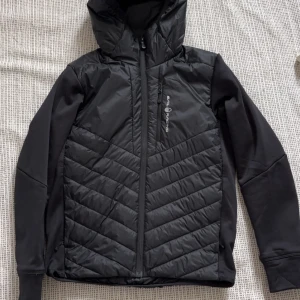 Sail racing cardigan - Snygg svart jacka från Sail racing med quiltad design och dragkedja framtill. Jackan har en huva och långa ärmar, perfekt för kyligare dagar. Den är stilren och funktionell, idealisk för både stad och natur.