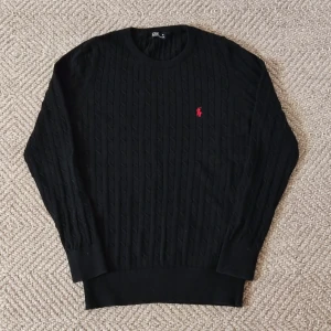 Svart stickad tröja från Ralph Lauren - Säljer en stilren svart stickad tröja från Ralph Lauren med kabelstickat mönster och röd broderad logotyp på bröstet. Tröjan har långa ärmar och rund halsringning. Perfekt för en klassisk look. Storlek XL i barnstorlek, passar som S/XS. Endast använd en gång.