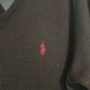 Svart tröja från Polo Ralph Lauren - Säljer en stilren svart tröja från Polo Ralph Lauren med v-ringning och lila broderad logga på bröstet. Tröjan har långa ärmar och är perfekt för en klassisk look.