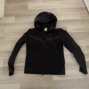 Svart Nike tech Fleece  - Säljer en svart hoodie från Nike med dragkedja och huva. Den har långa ärmar och en stilren design med diskreta detaljer. Perfekt för en casual look eller träning.