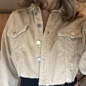 Beige croppad jacka från Zara - En beige croppad jacka från Zara TRF Collection. Jackan har ärmar, knappar framtill och två bröstfickor med knappar. Den är tillverkad i manchester och har en rå kant nedtill.