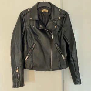 Svart skinnjacka från Glamorous - Snygg svart skinnjacka från Glamorous i storlek S. Jackan har en klassisk bikerstil med dragkedjor och knappar som detaljer. Perfekt för att ge din outfit en edgy touch.