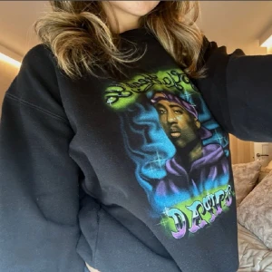 Svart sweatshirt med 2Pac-tryck  - Säljer en svart sweatshirt med ett stort färgglatt tryck av 2Pac på framsidan. Tröjan har långa ärmar och en rund halsringning. Perfekt för dig som gillar streetwear och vill ha en bekväm och stilren look.