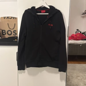 Svart hoodie från Hugo - Snygg svart hoodie från Hugo boss med dragkedja och röd Hugo logga på bröstet. Jättebra kvalite och andvänt ca 6-7 gånger. Säljer för att det är inte min stil längre. Orginalpris 1499kr men mitt pris (500kr) 
