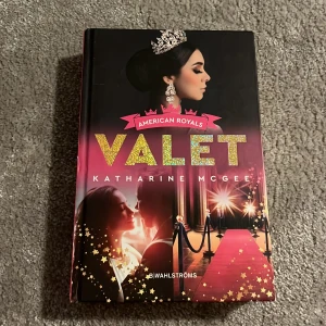 American Royals: Valet - Boken 'American Royals: Valet' av Katharine McGee utforskar en alternativ verklighet där USA styrs av en kungafamilj. Omslaget är färgstarkt med en bild av en kvinna med krona och en röd matta. Perfekt för dig som gillar intriger och kunglig dramatik.