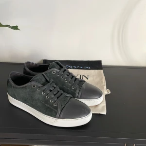 Gråa sneakers från Lanvin - Snygga gråa sneakers från Lanvin med svart tåhätta och snörning. Skorna har en stilren design med en vit sula som kontrasterar mot det gröna mockamaterialet. Perfekta för en avslappnad men elegant stil. Dustbag ingår! Skick 9-10 nästan helt nya bara några repor på captoen! Sjukt bra deal. Vid frågor är det bara att höra av sig.