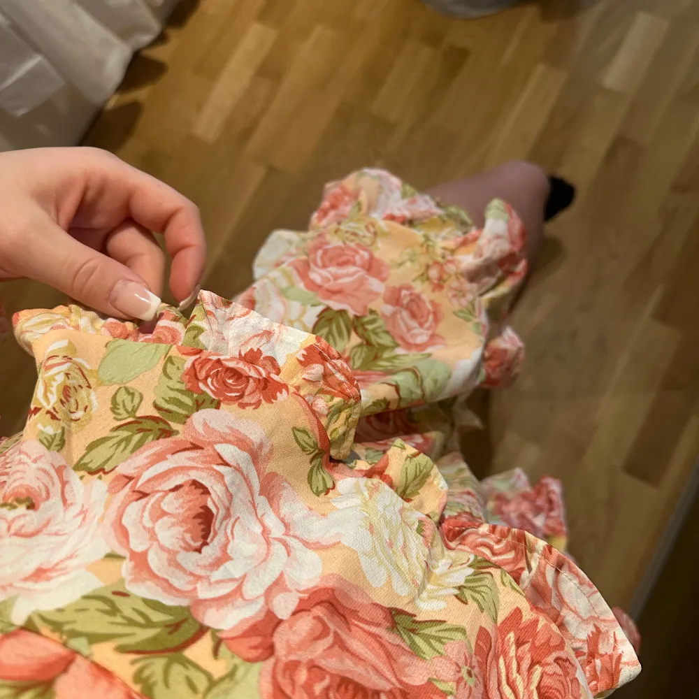 Charmig blommig klänning med volanger och v-ringad design. Klänningen har långa ärmar och ett knytband i midjan för en smickrande passform. Perfekt för somriga dagar med sitt färgglada mönster i rosa och grönt.. Mekot.