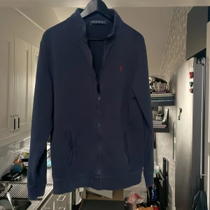 Mörkblå tröja med dragkedja från Polo Club - Säljer en stilren mörkblå tröja från Polo Club med dragkedja och broderad röd logotyp på bröstet. Tröjan har långa ärmar och en hög krage, perfekt för en avslappnad look. Passar bra till både jeans och chinos.