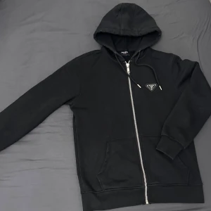 Svart hoodie pr*da - Säljer en stilren svart hoodie med dragkedja och huva. Den har en diskret logga på bröstet och ribbade muddar vid ärmslut och nederkant. Perfekt för en casual look.