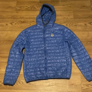 Blå quiltad jacka med huva - Säljer en blå quiltad jacka med huva och dragkedja framtill. Jackan har ett märke på bröstet och är perfekt för kyligare dagar. Den är lätt och smidig att bära med sig.